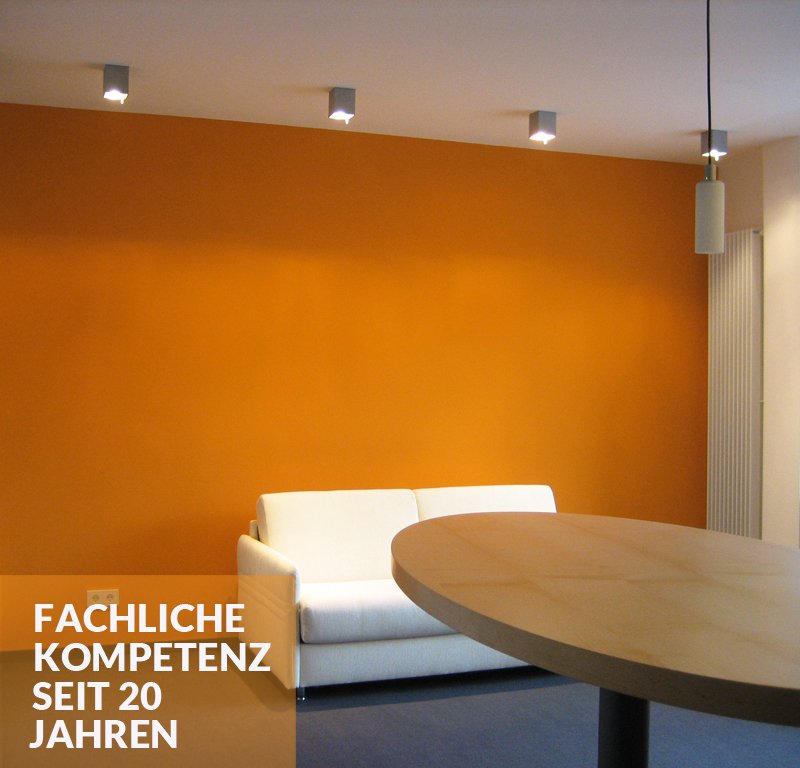 Fachliche Kompetenz seit 20 Jahren - Blick in ein Wohnzimmer. Im Vordergrund befindet sich ein runder Holztisch, im Hintergrund ein schlichtes, weißes Sofa vor orangener Wand.