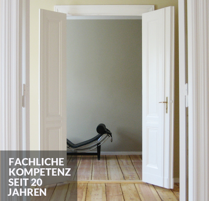 Fachliche Kompetenz seit 20 Jahren - Blick durch zwei geöffnete, weiße zweiblättrige Türen auf ein schwarzes modernes Sofa vor heller Wand auf rustikalem Dielenboden.